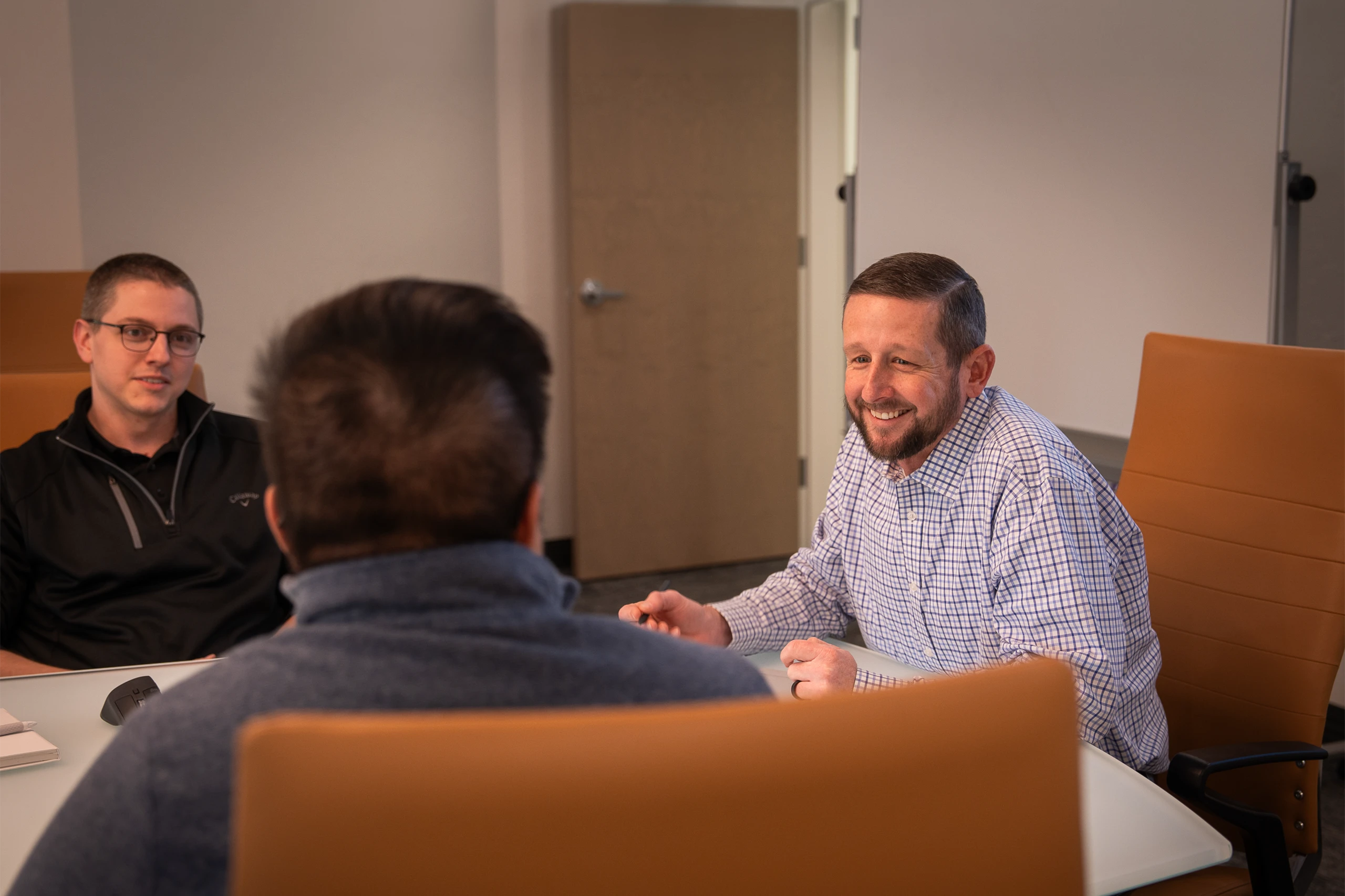 Three smiling men sitting around a table talking.