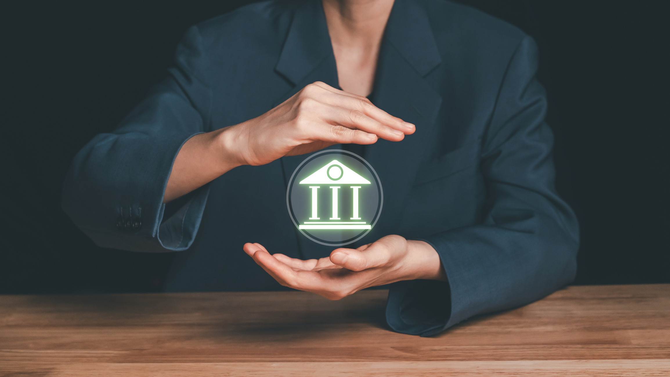 Businesswoman hands covering glowing bank building icon, symbolizing financial institution protection, banking system stability, and regulatory compliance.